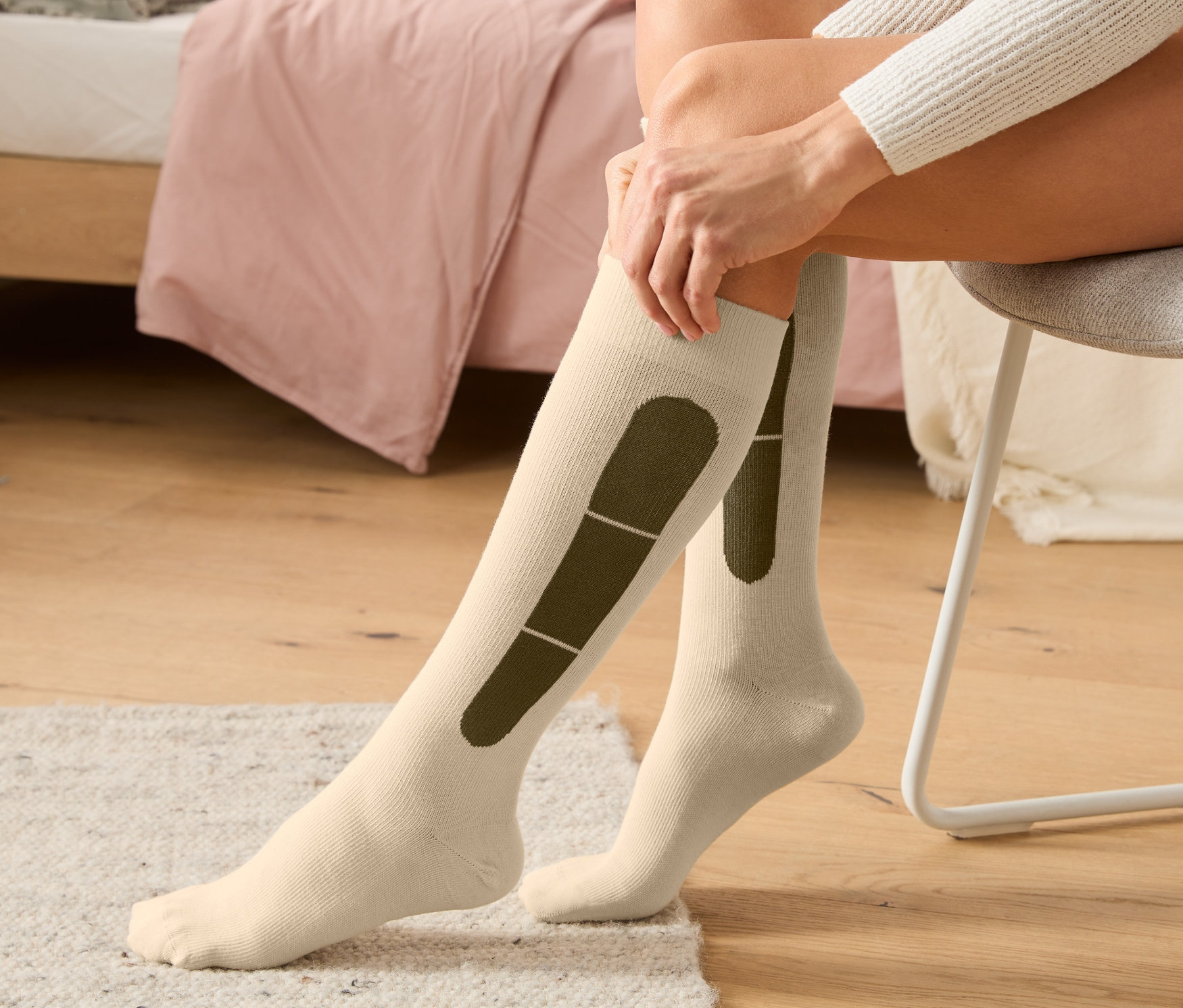 Frau sitzt auf einem Stuhl und zieht beige Reise-Kompressionsstrümpfe an.