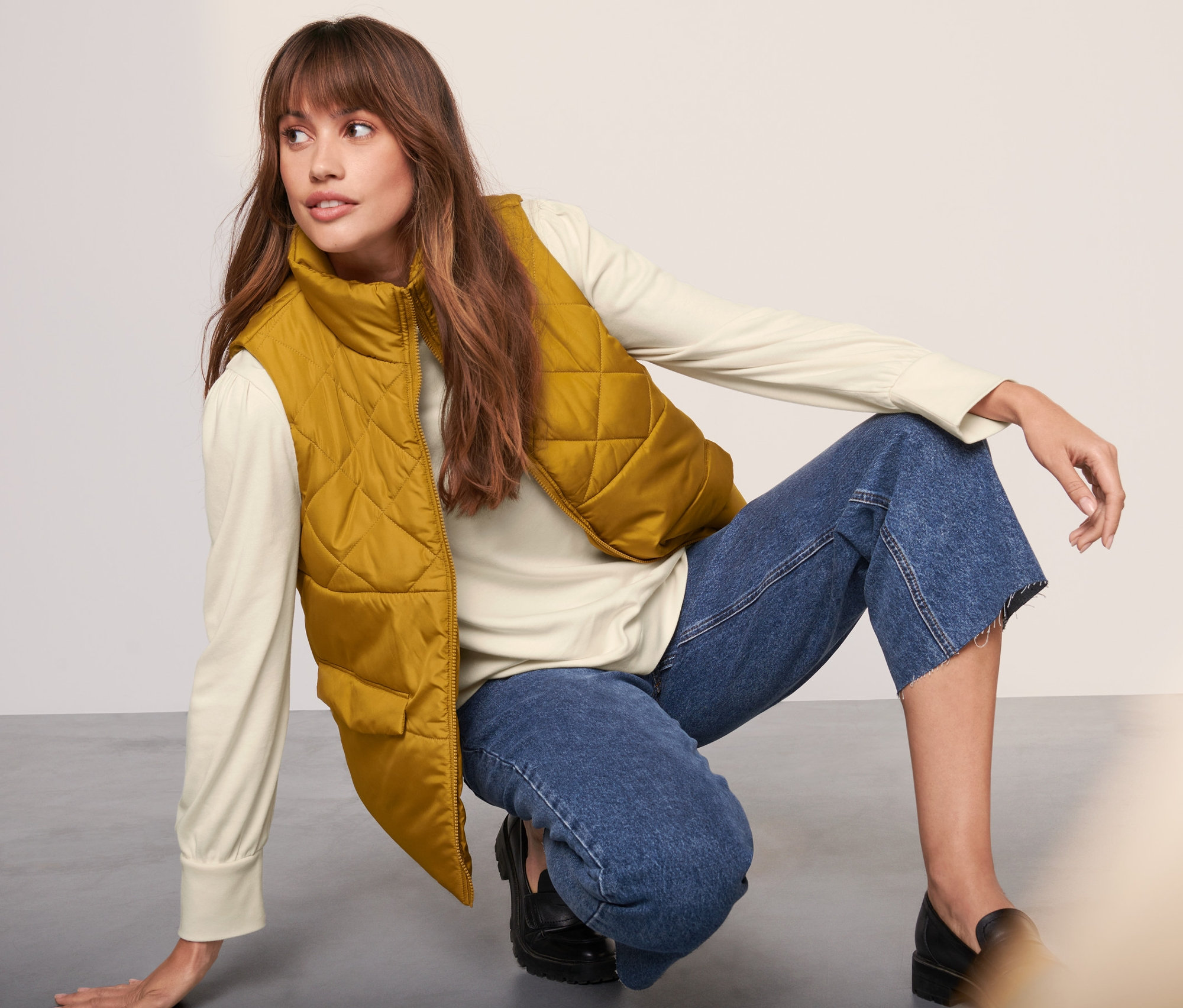 Frau in Steppweste, Pullover und Jeans posiert auf dem Boden.