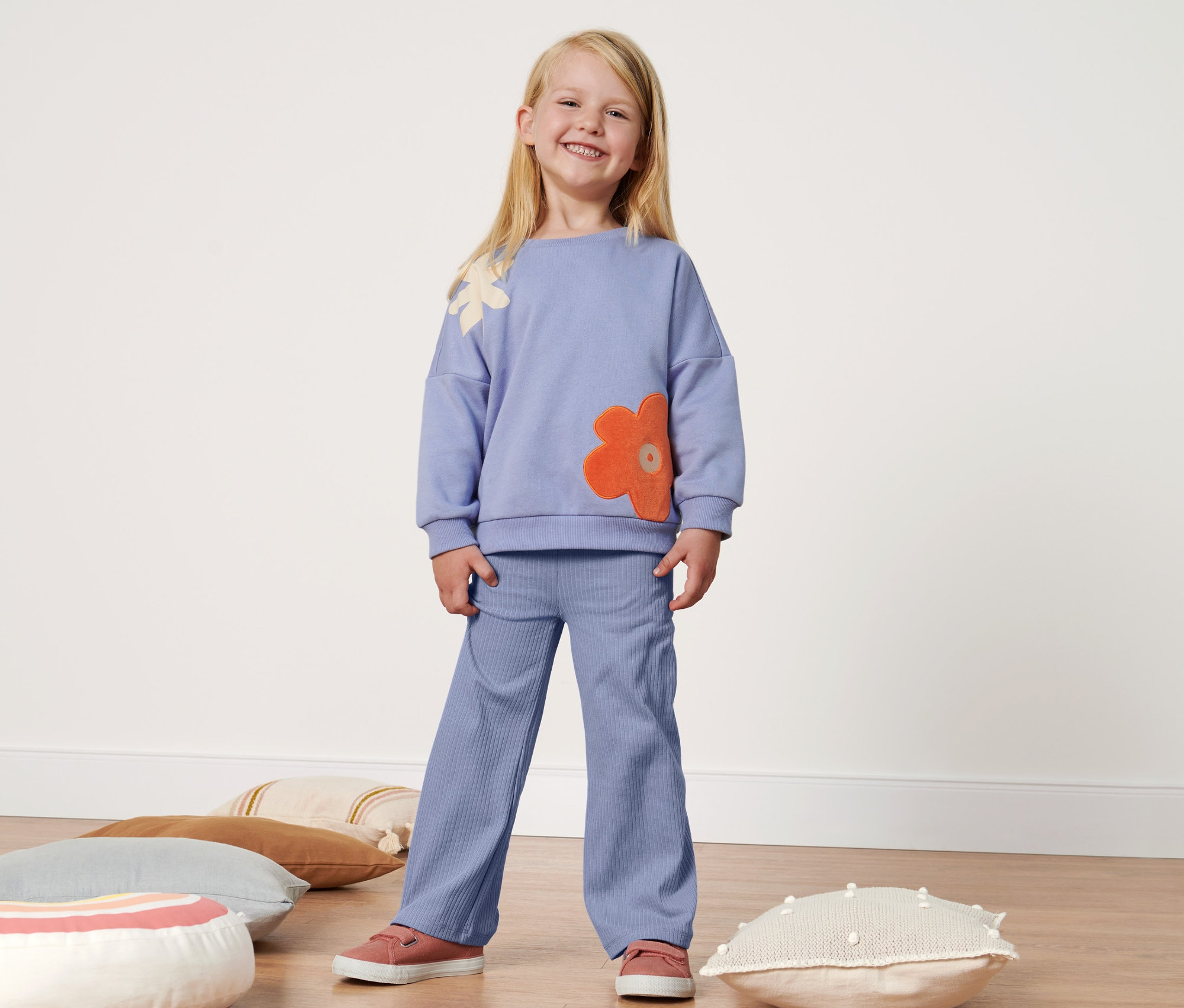 Lächelndes blondes Mädchen steht und trägt ein lila Outfit mit Blumen und eine Kleinkind-Rippenleggings mit Schlag.