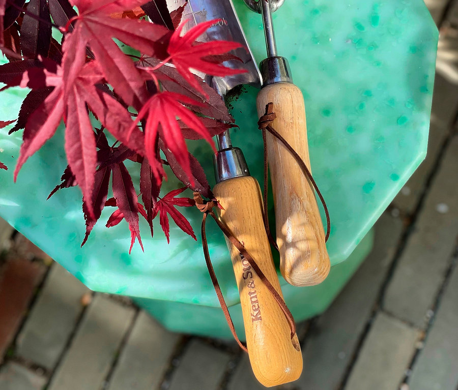 Zwei Gartenkellen mit Holzgriffen liegen auf einem grünen Beistelltisch neben roten Blättern.