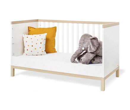 Ein Kinderbett mit weißem Rahmen und Holzfüßen. Im Bett liegen ein grauer Plüsch-Elefant und zwei Kissen.