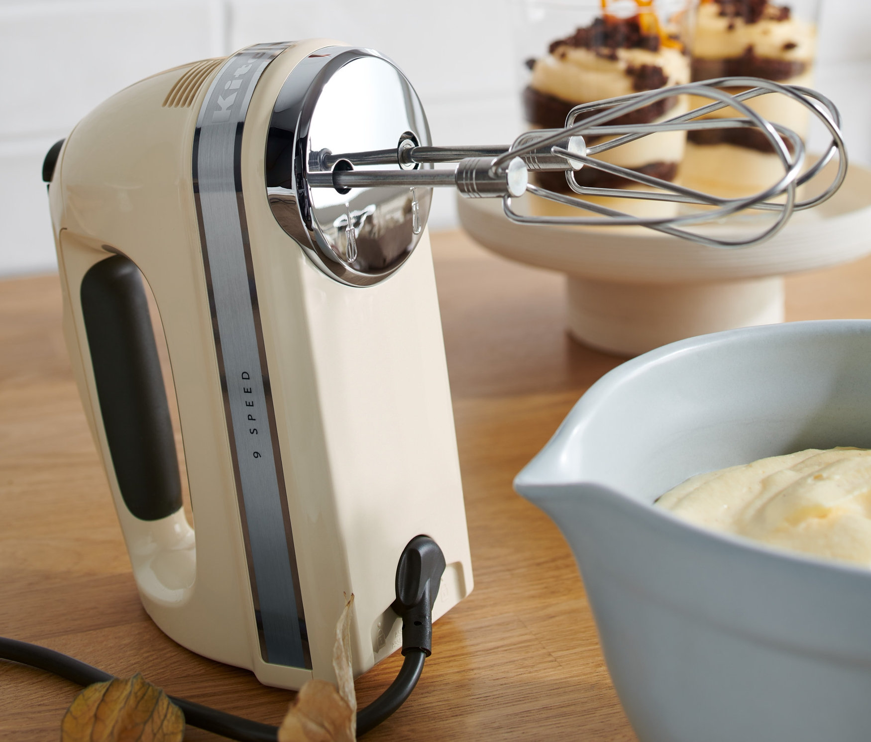 Ein cremefarbener KitchenAid-Handmixer »5KHM9212EAC« steht neben einer Schüssel Teig und zwei Desserts.