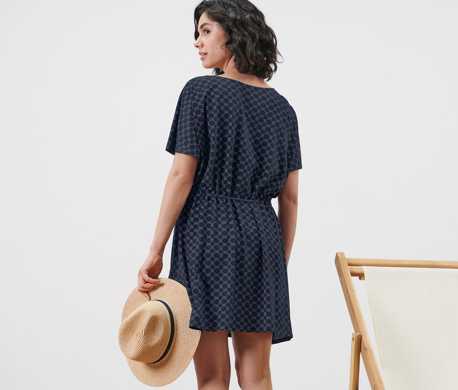 Frau im Strandtunika hält Hut neben einem Liegestuhl.