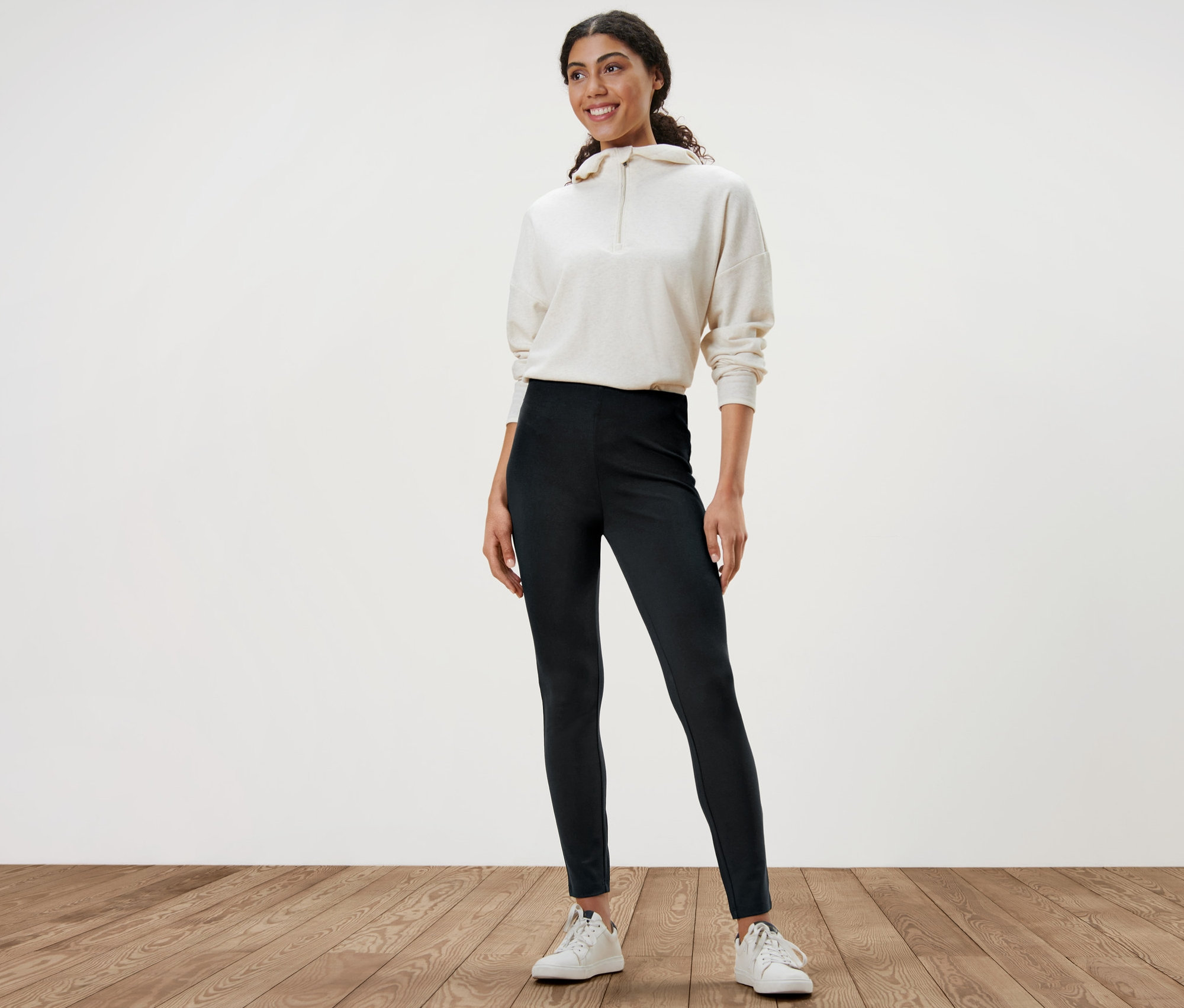 Frau steht und trägt schwere Leggings, einen weißen Pullover und weiße Turnschuhe.