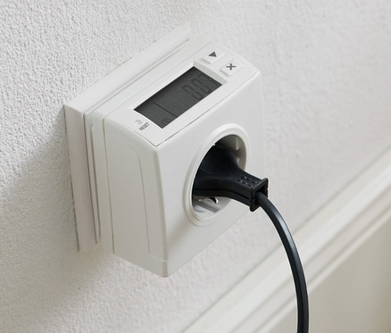 An einer weißen Wand befindet sich eine Steckdose mit einem Energieverbrauchsmessgerät und einem eingesteckten schwarzen Kabel.