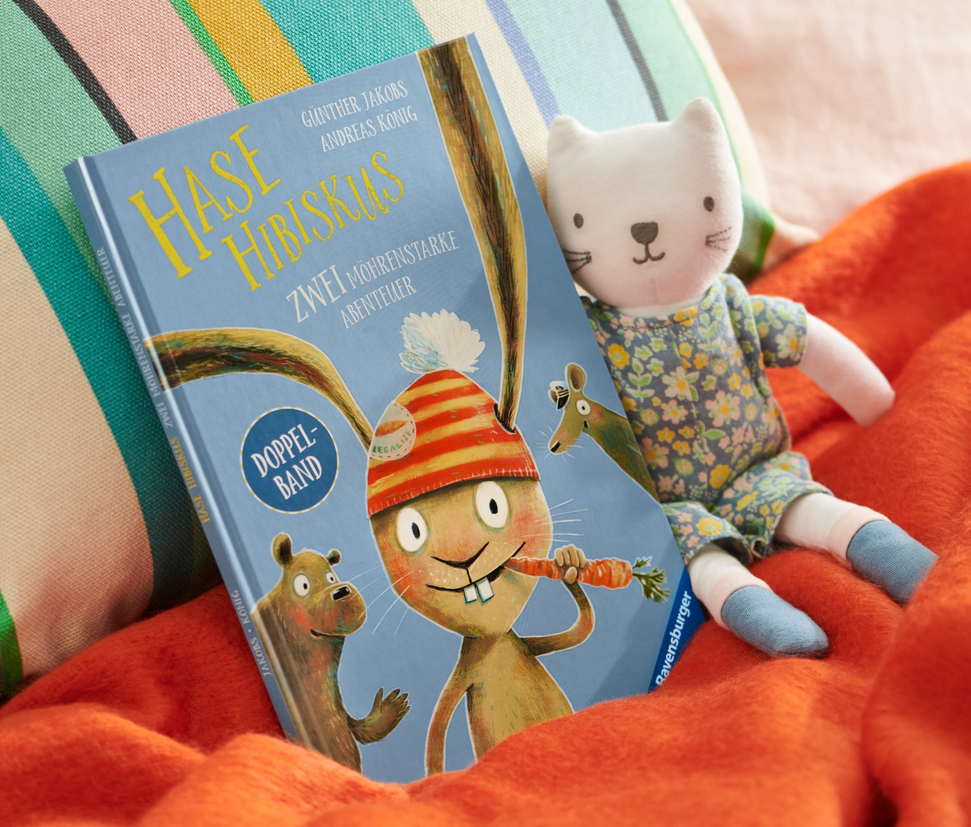 Auf einer orangefarbenen Decke liegen ein »Hase Hibiskus« Buch, daneben ein Katzenstofftier und dahinter ein buntes Kissen.