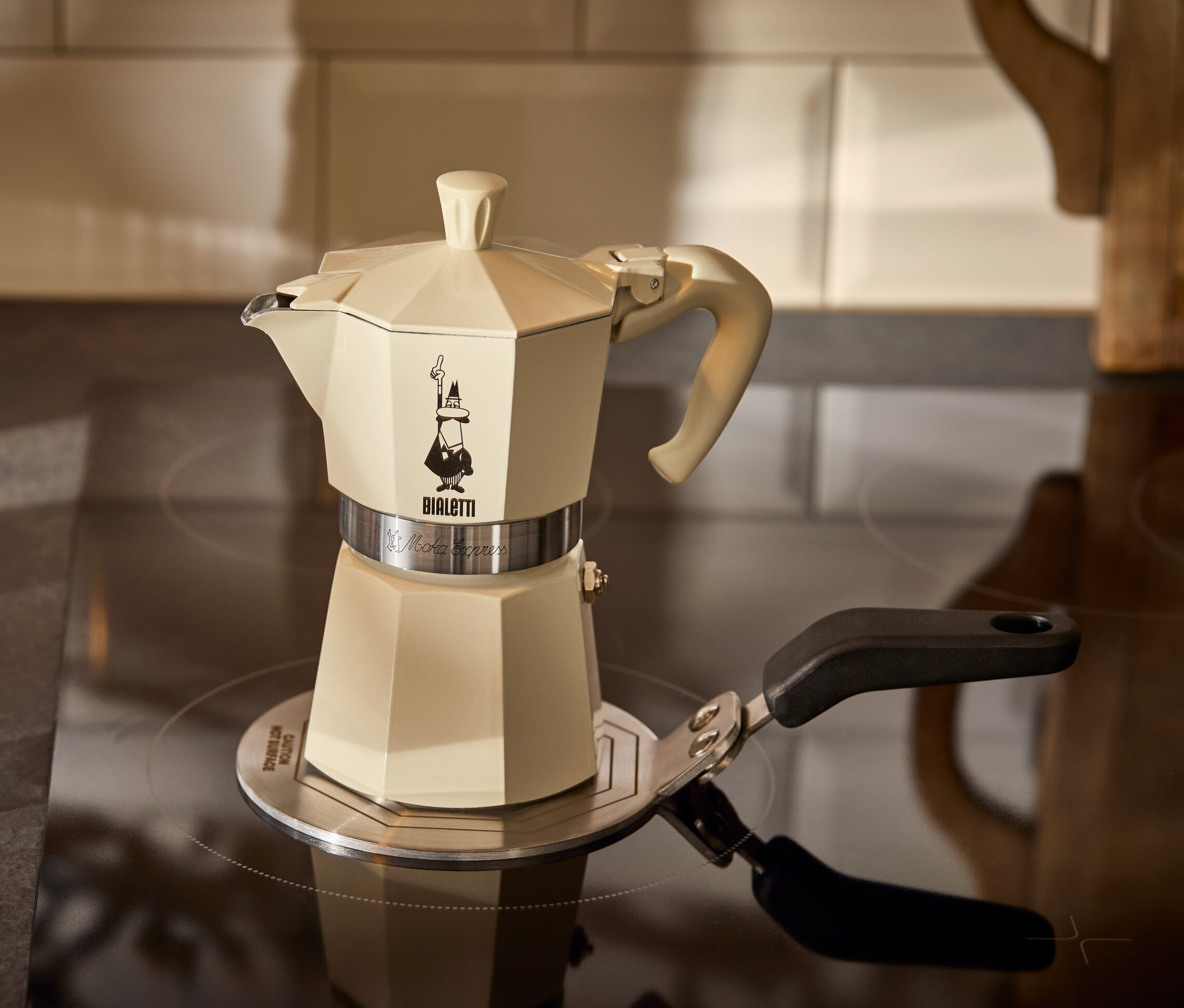 Ein cremefarbener Espressokocher Bialetti Moka Express 190 ml steht auf einer Bialetti Adapterplatte Induktion 13 cm neben einem Glas Espresso.