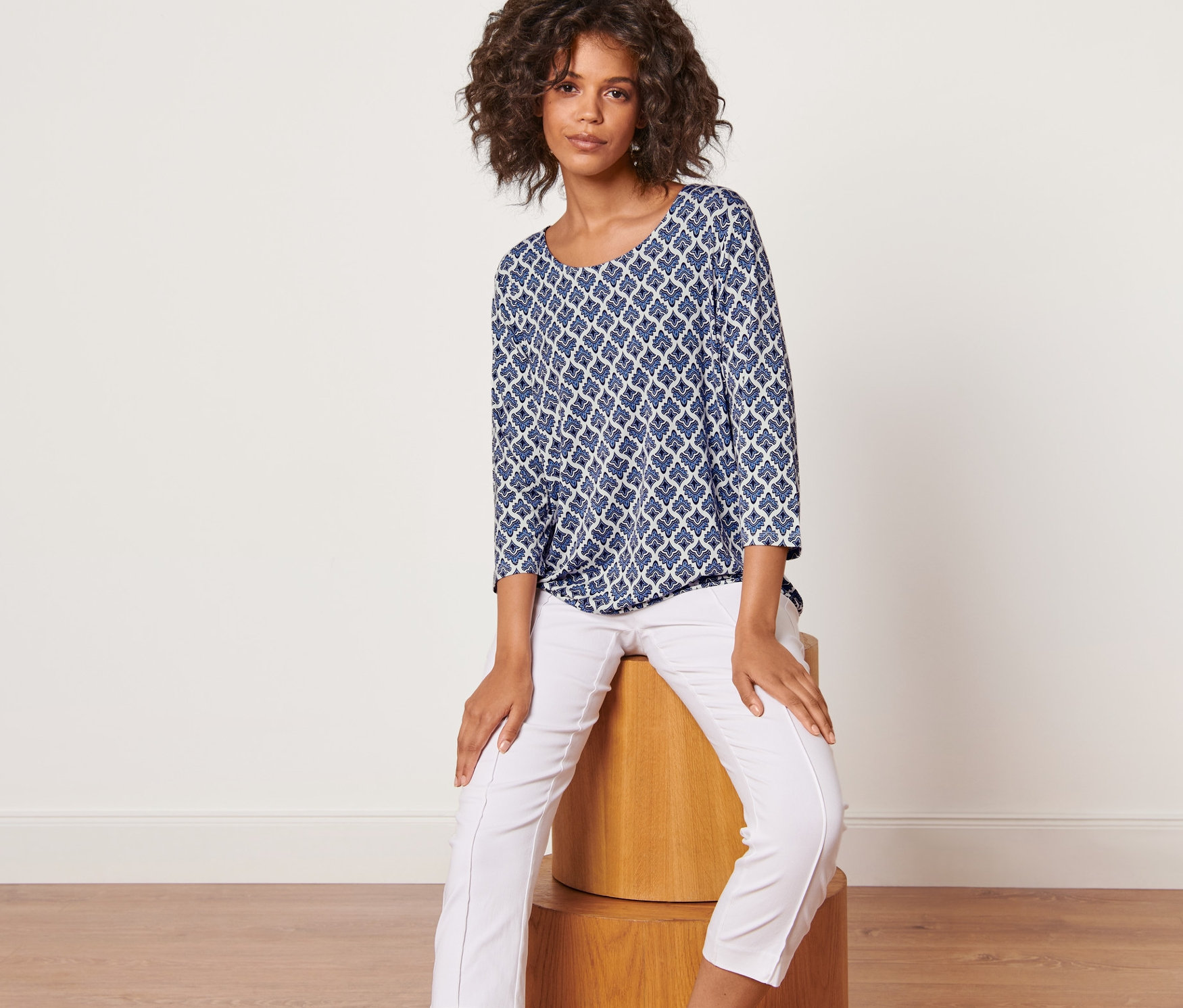 Frau sitzt auf einem Holzhocker und trägt ein Blusenshirt mit Alloverprint und eine weisse Hose.