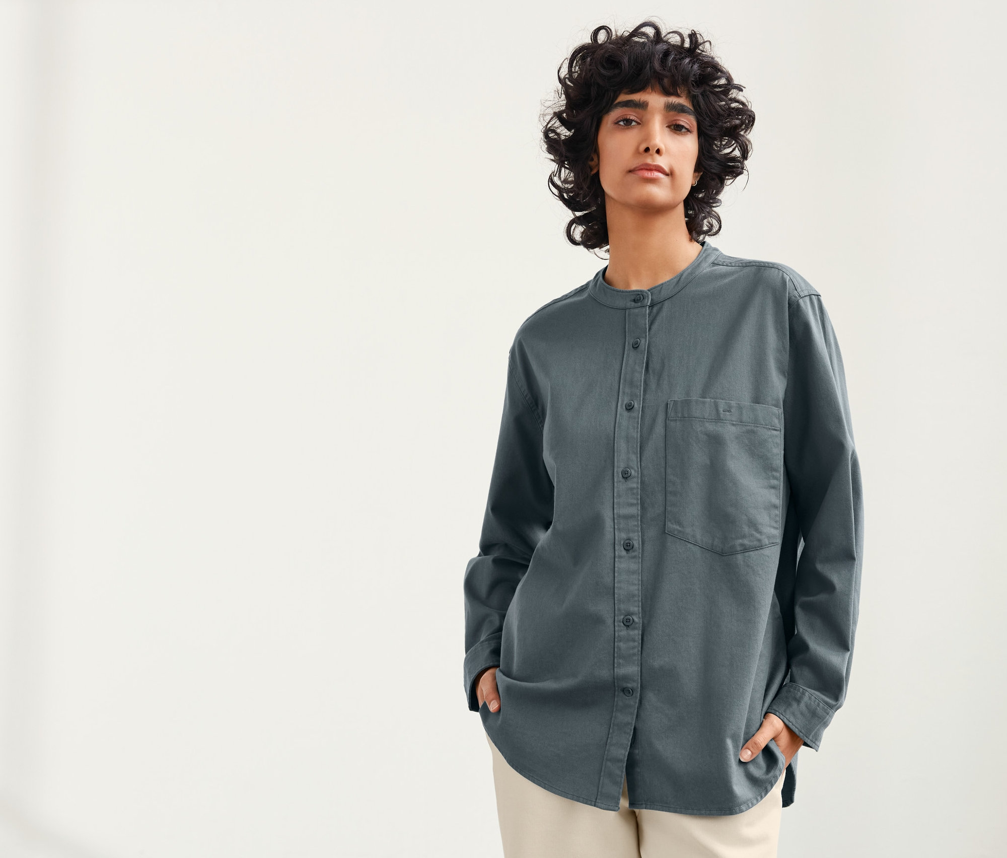 Frau mit dunklen lockigen Haaren posiert vor hellem Hintergrund und trägt eine blau-graue Leinenbluse von NAH/STUDIO.