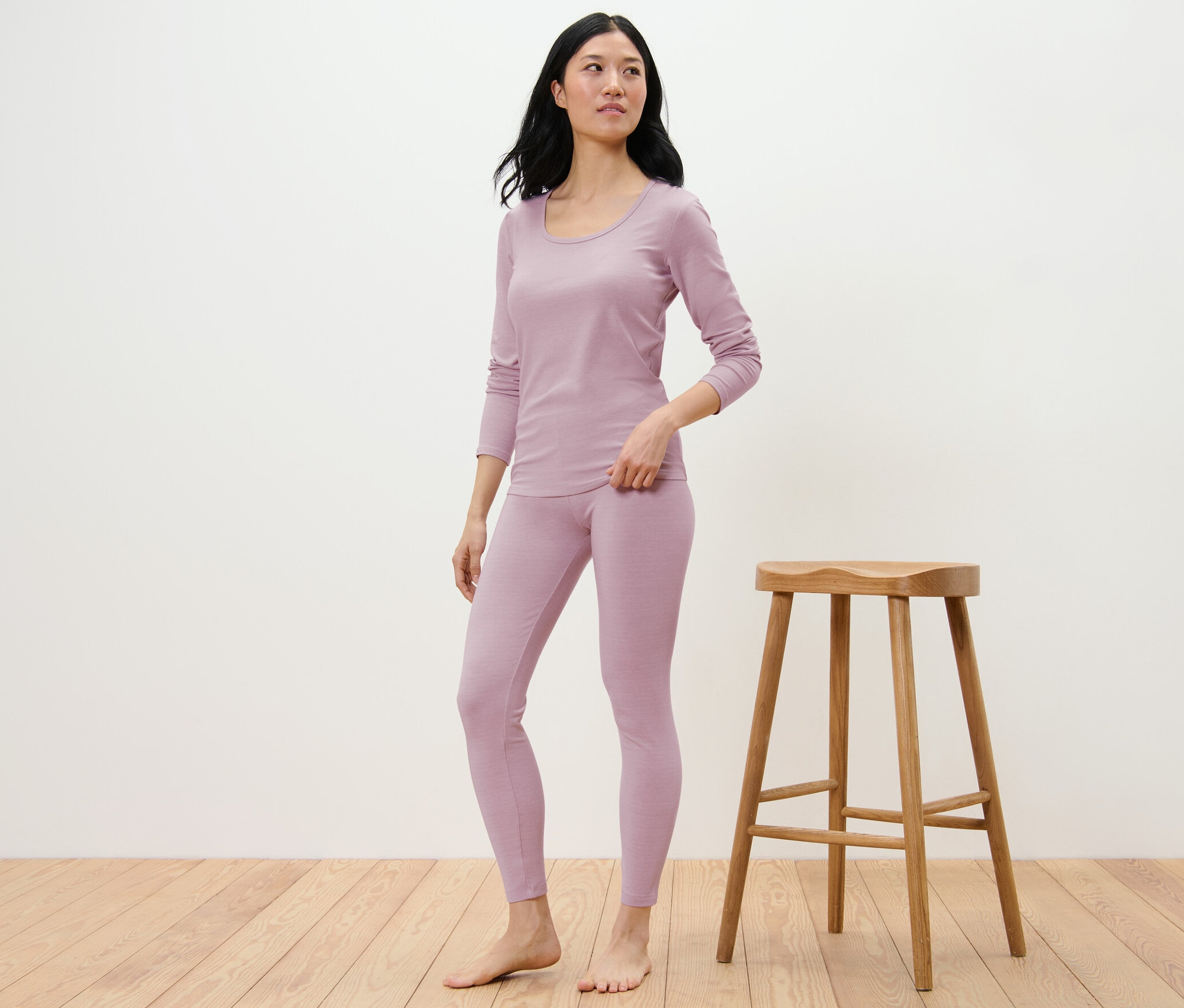 Frau posiert in lila farbenen Leggings mit RWS-zertifizierter Wolle und langärmligem Oberteil neben einem Holzhocker.