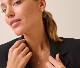 Blonde Frau im schwarzen Sakko berührt Revers und trägt goldene Creolen, Ring und zwei Halsketten, eine davon mit Kreuz. Sie trägt eine 375 Gold Creolen Zirkonia, 375 Gold Ring Eternity Zirkonia, 375 Gold Kette Singapur, 375 Gold Kette Kreuz, 585 Gold Kette Kreuz und 585 Gold Ring Eternity Zirkonia.