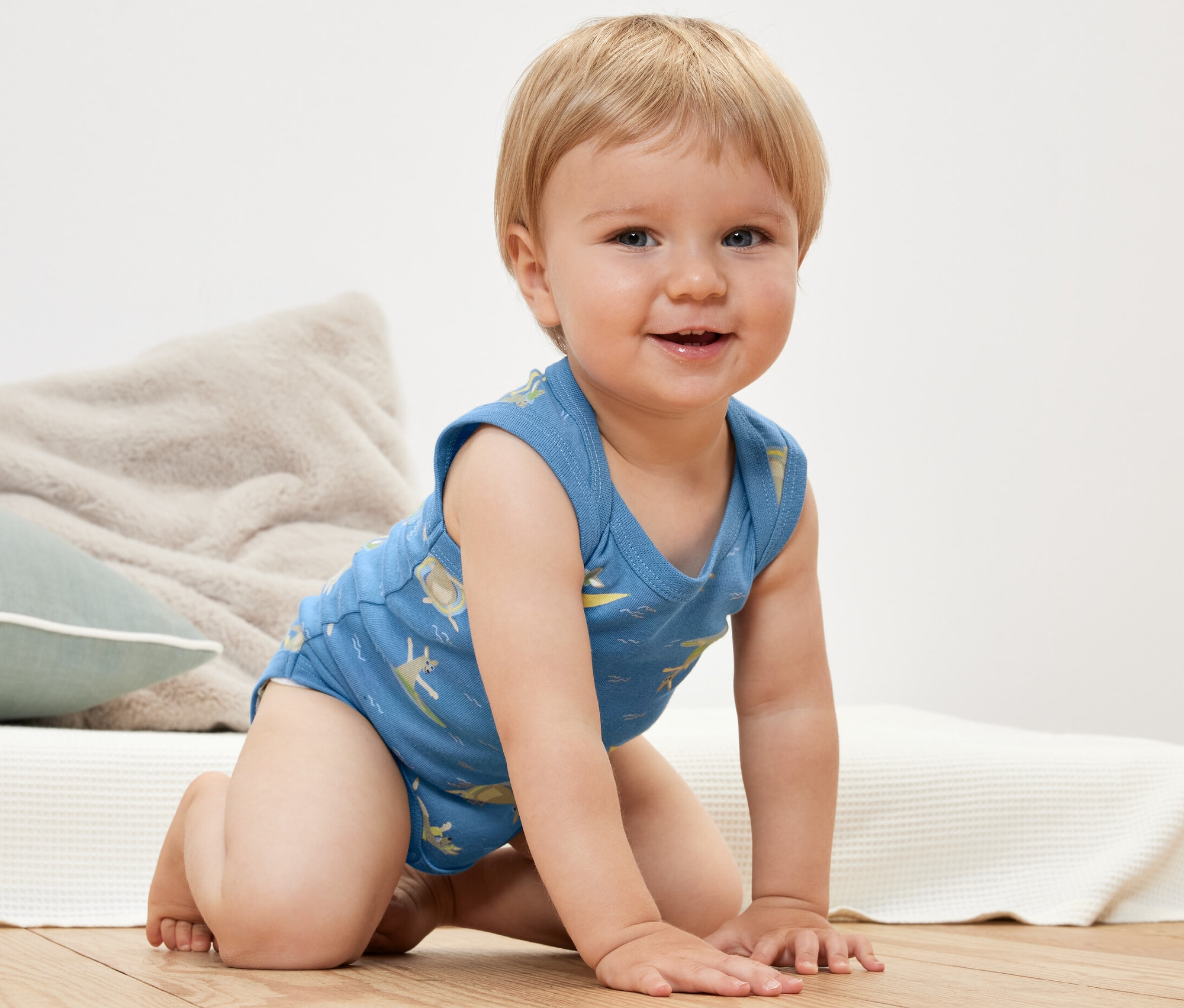 Lächelnder Säugling kniet und trägt einen blauen 2 Baby-Bodys mit Aufdruck.