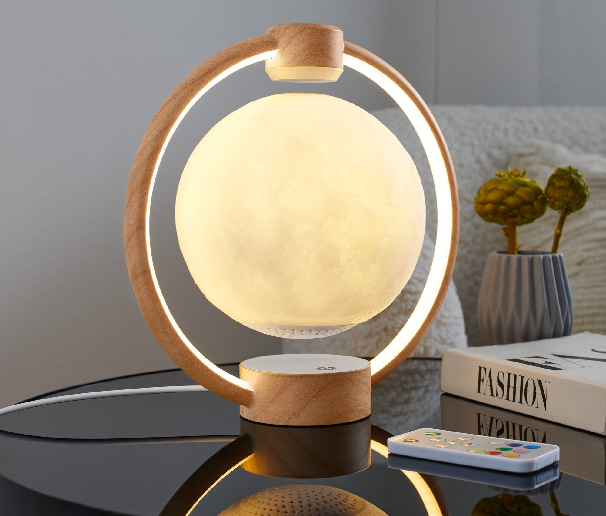 Schwebender LED-Bluetooth®-Lautsprecher »Mond« steht auf einem Nachttisch neben einem Buch und einer Fernbedienung.