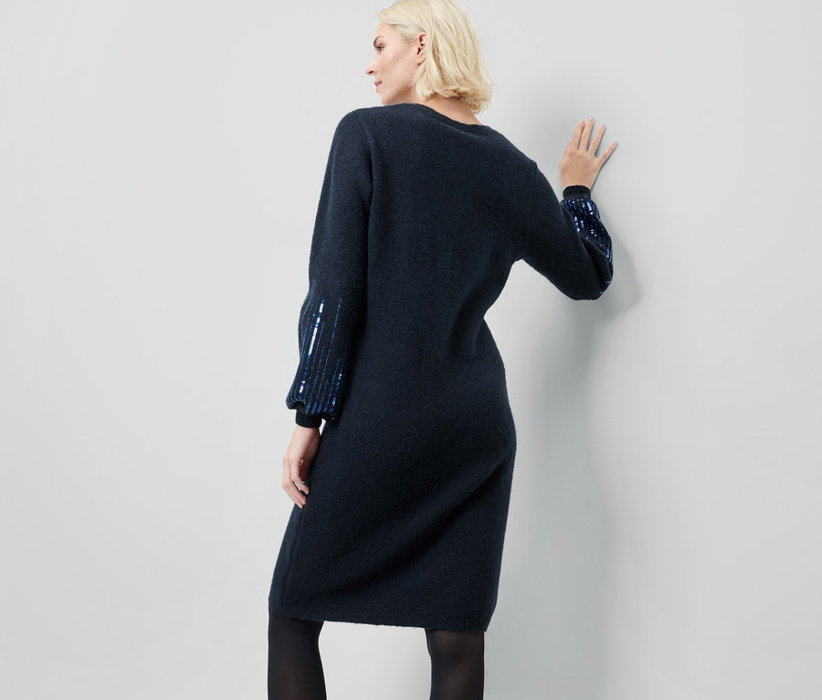 Frau mit blonden Haaren posiert mit dem Rücken zur Kamera und trägt ein dunkelblaues Strickkleid mit Pailletten.