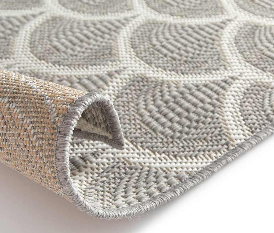 Nahaufnahme eines beigen Outdoor-Teppichs »Arcs« mit geometrischem Muster und eingerollter Ecke.