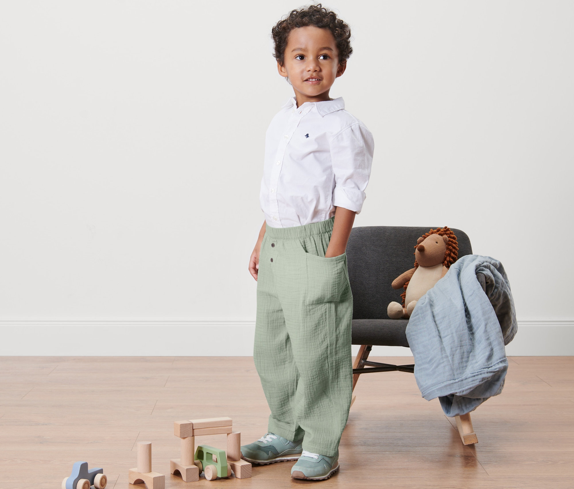 Ein Junge steht mit den Händen in den Taschen einer Kinder-Premium-Hose. Neben ihm stehen ein grauer Sessel mit einem Spielzeug und Holzspielzeug.