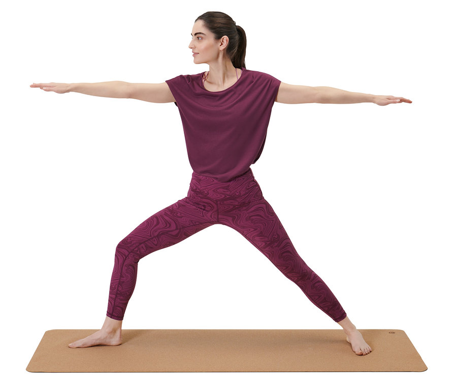 Frau in lila Sportbekleidung macht Krieger-II-Pose auf Kork-Yoga-und-Fitnessmatte.