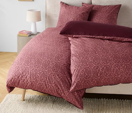 Bettwäsche mit Blumenmuster in Rosa- und Burgundertönen auf einem Bett mit beigem Kopfteil.