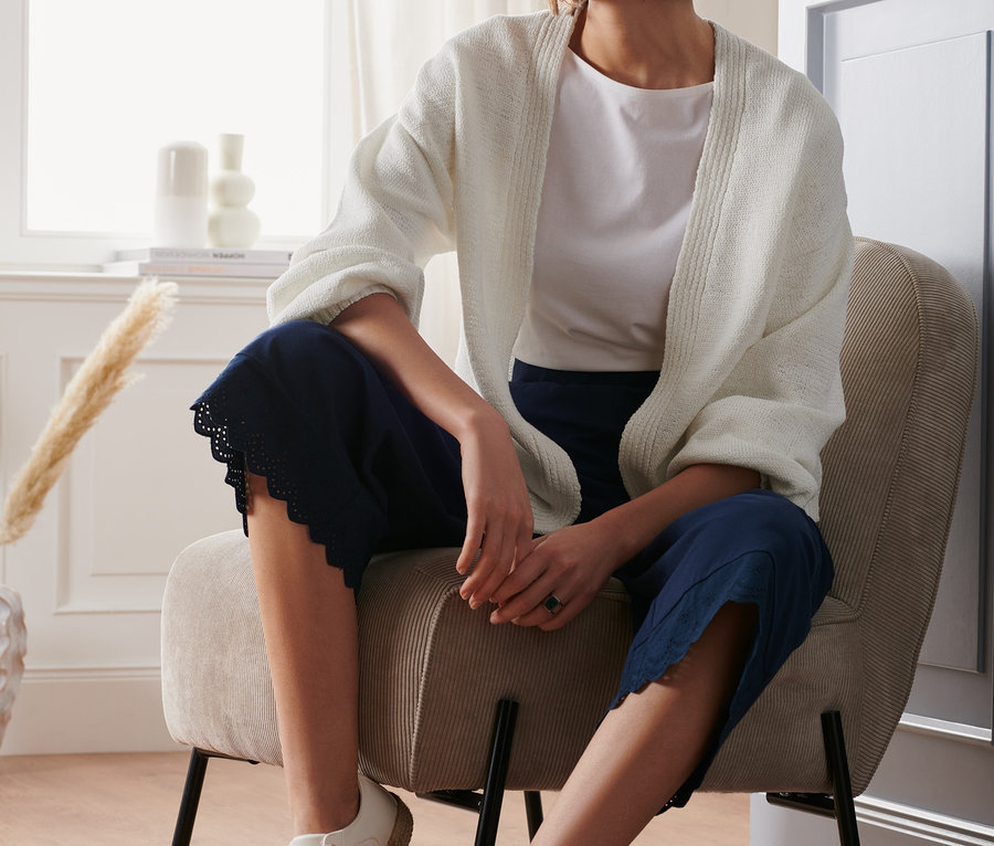 Frau sitzt im Sessel und trägt einen offenen Cardigan aus Bändchengarn, ein weißes T-Shirt und eine blaue Hose.