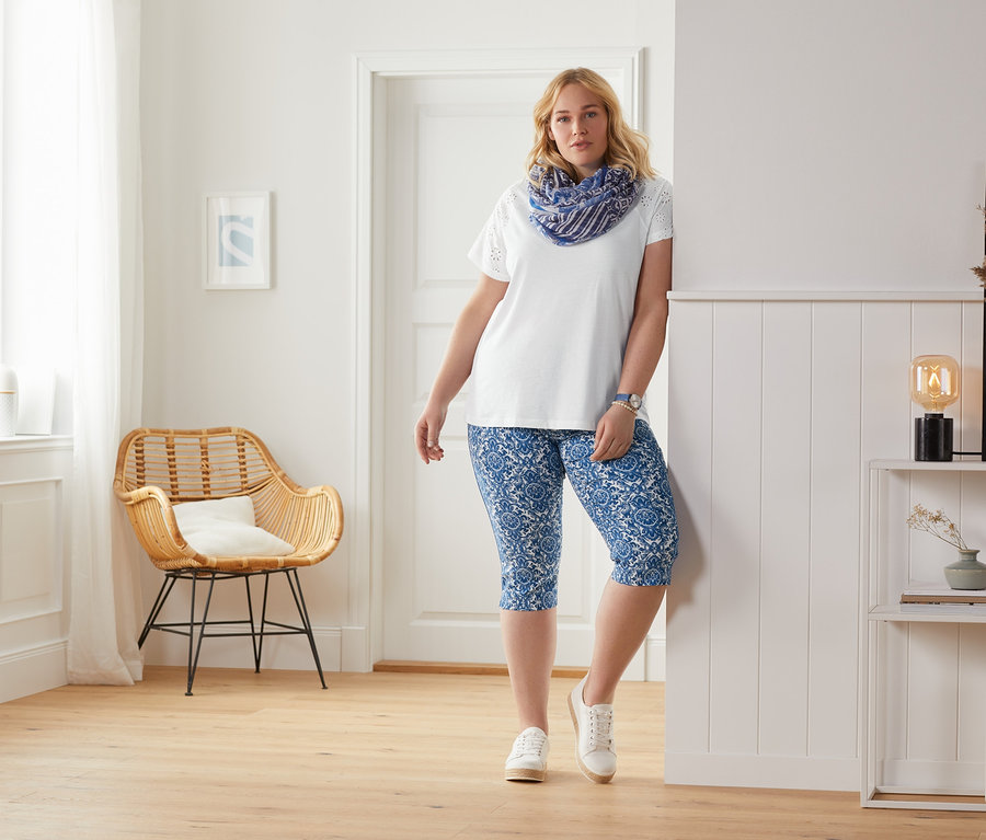 Eine Frau lehnt an der Wand und trägt ein weißes Shirt mit Stickerei und eine blaue, bedruckte Stretchhose in 3/4-Länge.