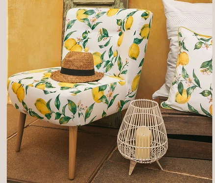 Auf einem Cocktailsessel »Fiona« mit Zitronenprint liegt ein Hut. Neben dem Sessel stehen Perkal-Bettwäsche mit Stickerei, Normalgröße, und ein Wende-Dekokissenbezug. Im Hintergrund ist eine gelbe Wand zu sehen. Ein Jumbobecher steht auch da.