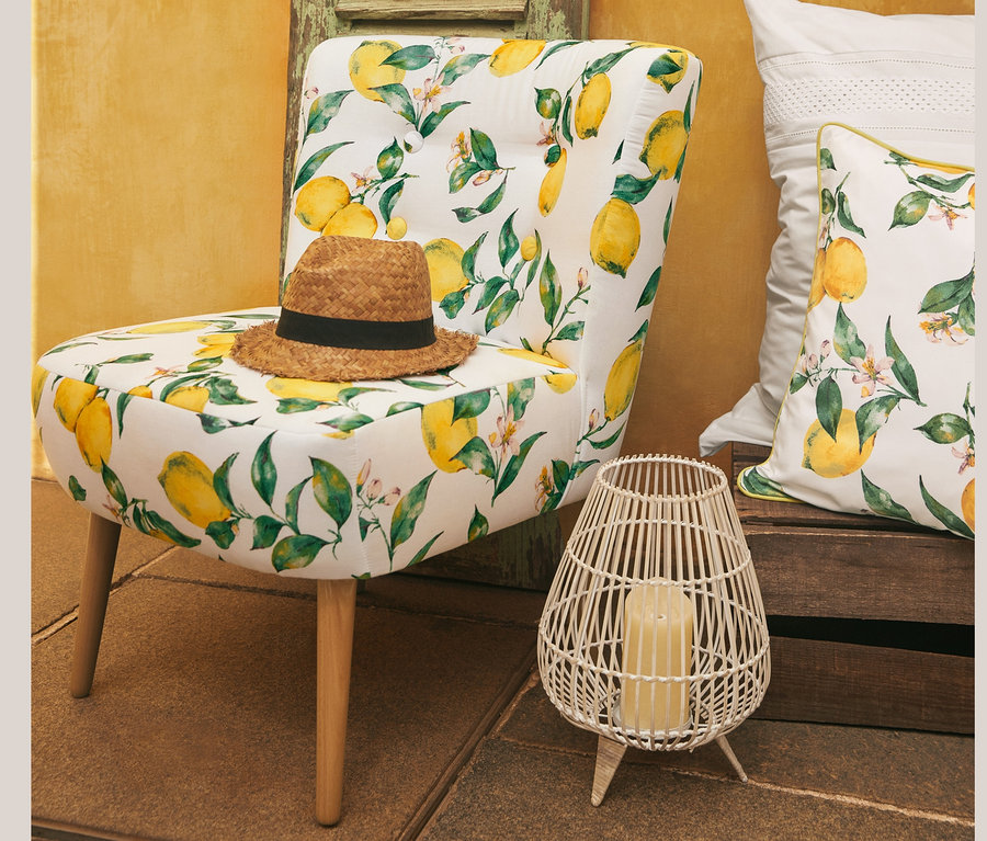 Auf einem Cocktailsessel »Fiona« mit Zitronenprint liegt ein Hut. Neben dem Sessel stehen Perkal-Bettwäsche mit Stickerei, Normalgröße, und ein Wende-Dekokissenbezug. Im Hintergrund ist eine gelbe Wand zu sehen. Ein Jumbobecher steht auch da.