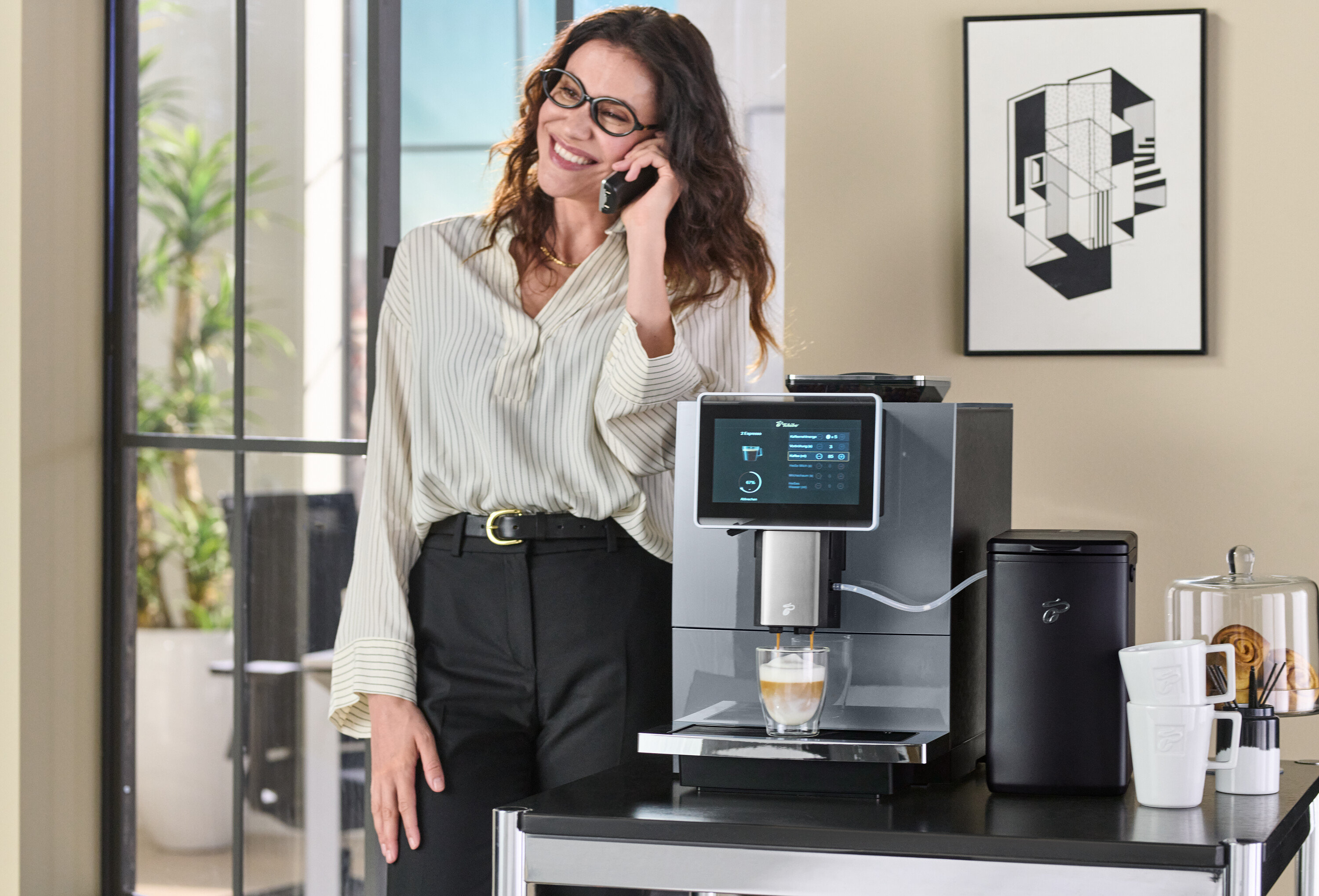 Frau telefoniert und lehnt sich an einen Kaffeevollautomaten, aus dem Kaffee in ein Glas läuft.