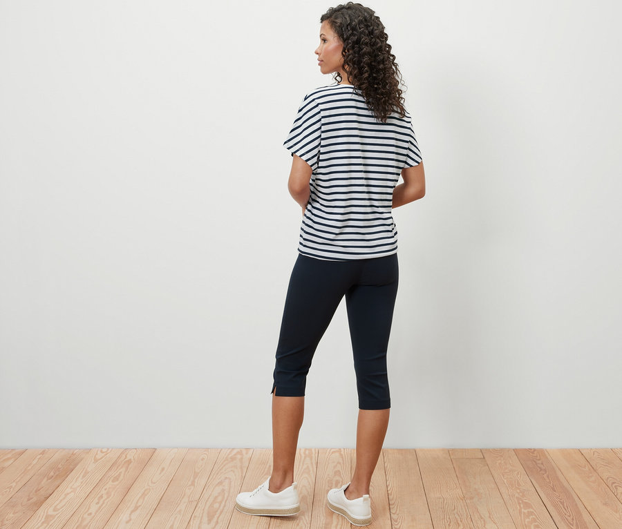 Eine Frau steht mit dem Rücken zur Kamera und trägt ein überschnittenes Blusenshirt mit Streifen, eine dunkelblaue Dreiviertelhose und weiße Turnschuhe.