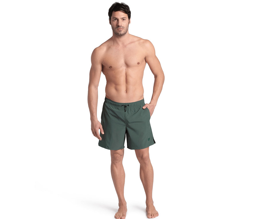 Mann steht oberkörperfrei und trägt grüne arena Evo Badeshorts, eine Hand in der Tasche.
