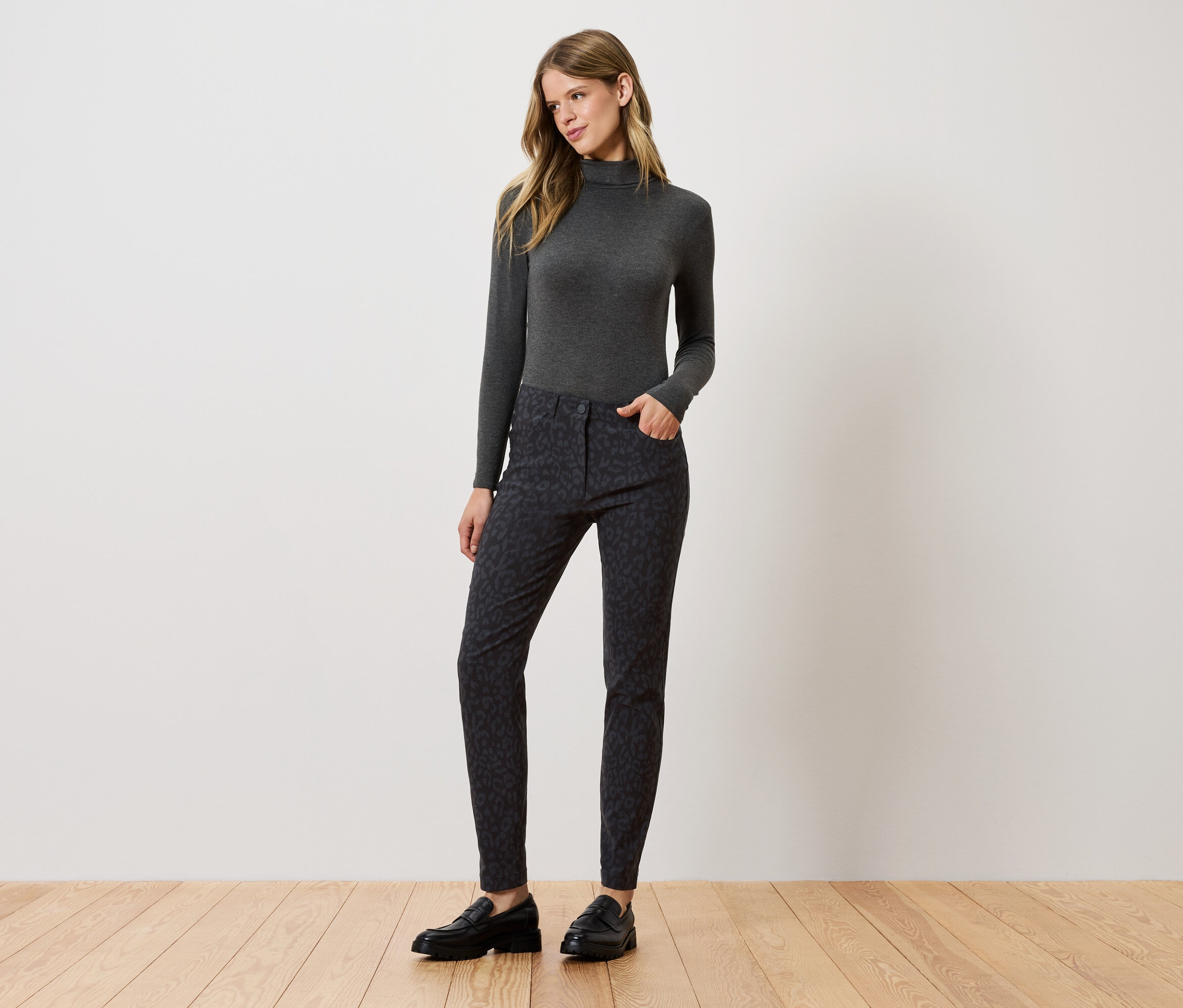 Eine Frau steht und posiert in einem grauen Rollkragenpullover, gemusterten Hosen und schwarzen Schuhen.