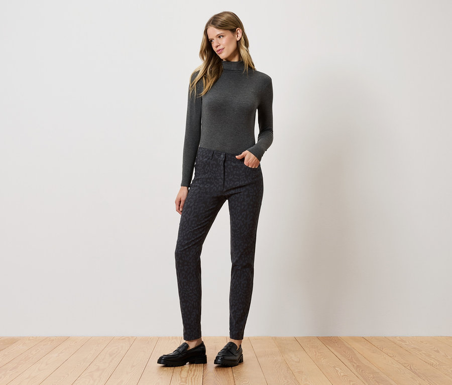 Eine Frau steht und posiert in einem grauen Rollkragenpullover, gemusterten Hosen und schwarzen Schuhen.