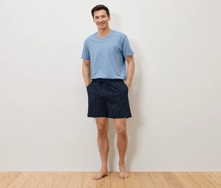 Ein Mann steht barfuß mit den Händen in den Taschen seiner marineblauen, gepunkteten Shorts da und trägt ein hellblaues T-Shirt vor einer weißen Wand.