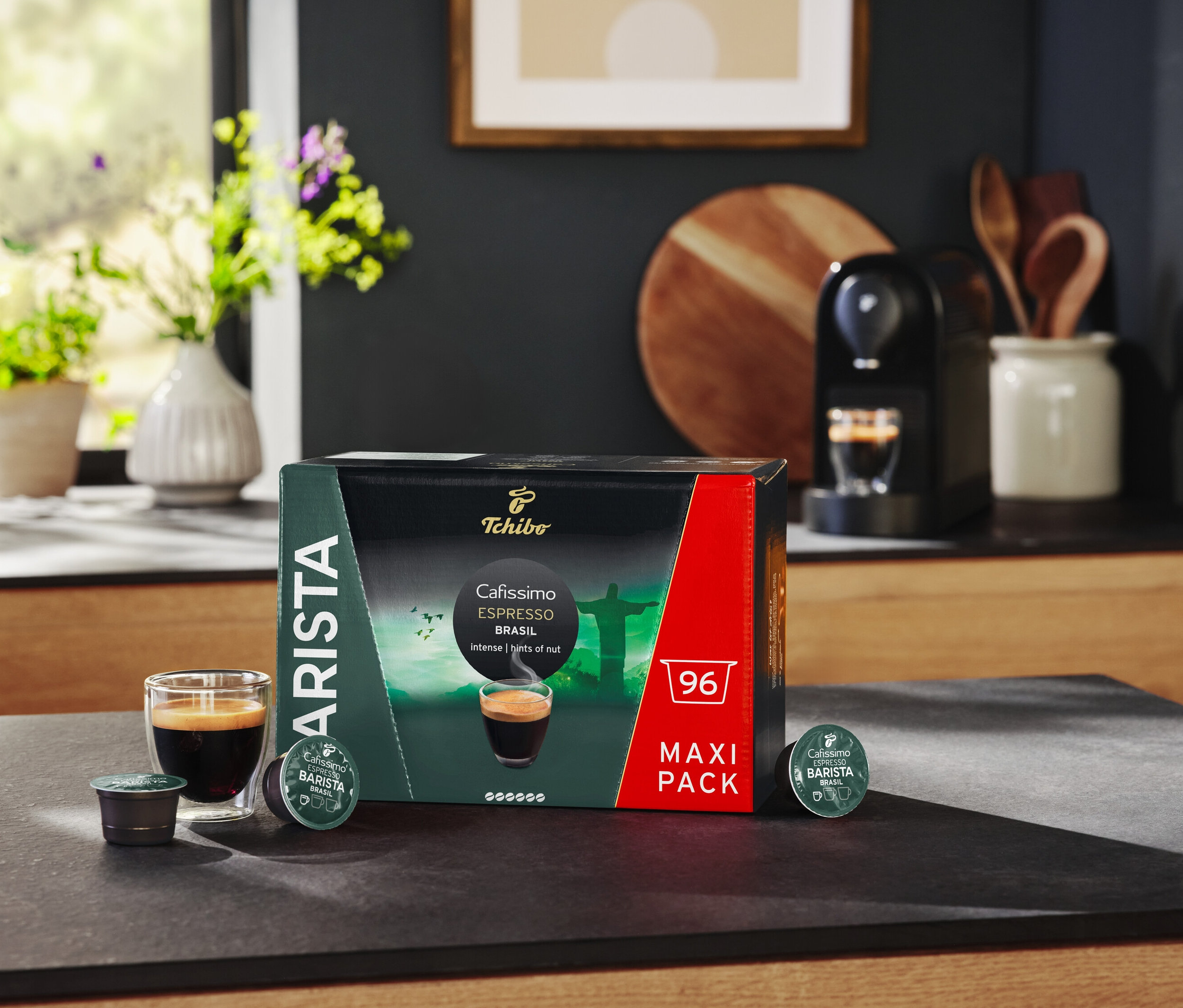 Auf einem Tisch stehen eine Packung Cafissimo BARISTA Espresso Brasil - 96 Kapsel, ein Glas Espresso und eine Kaffeemaschine.