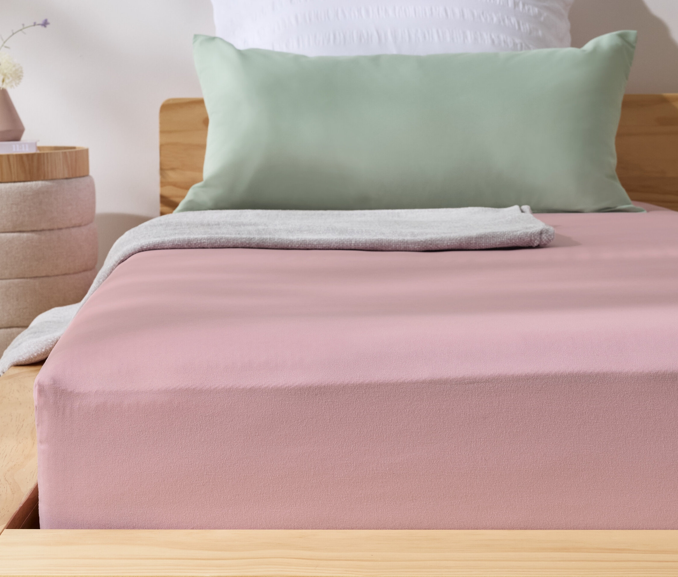 Rosa Microfaser-Spannbettlaken auf einem Bett. Daneben steht ein Nachttisch mit einer Vase.