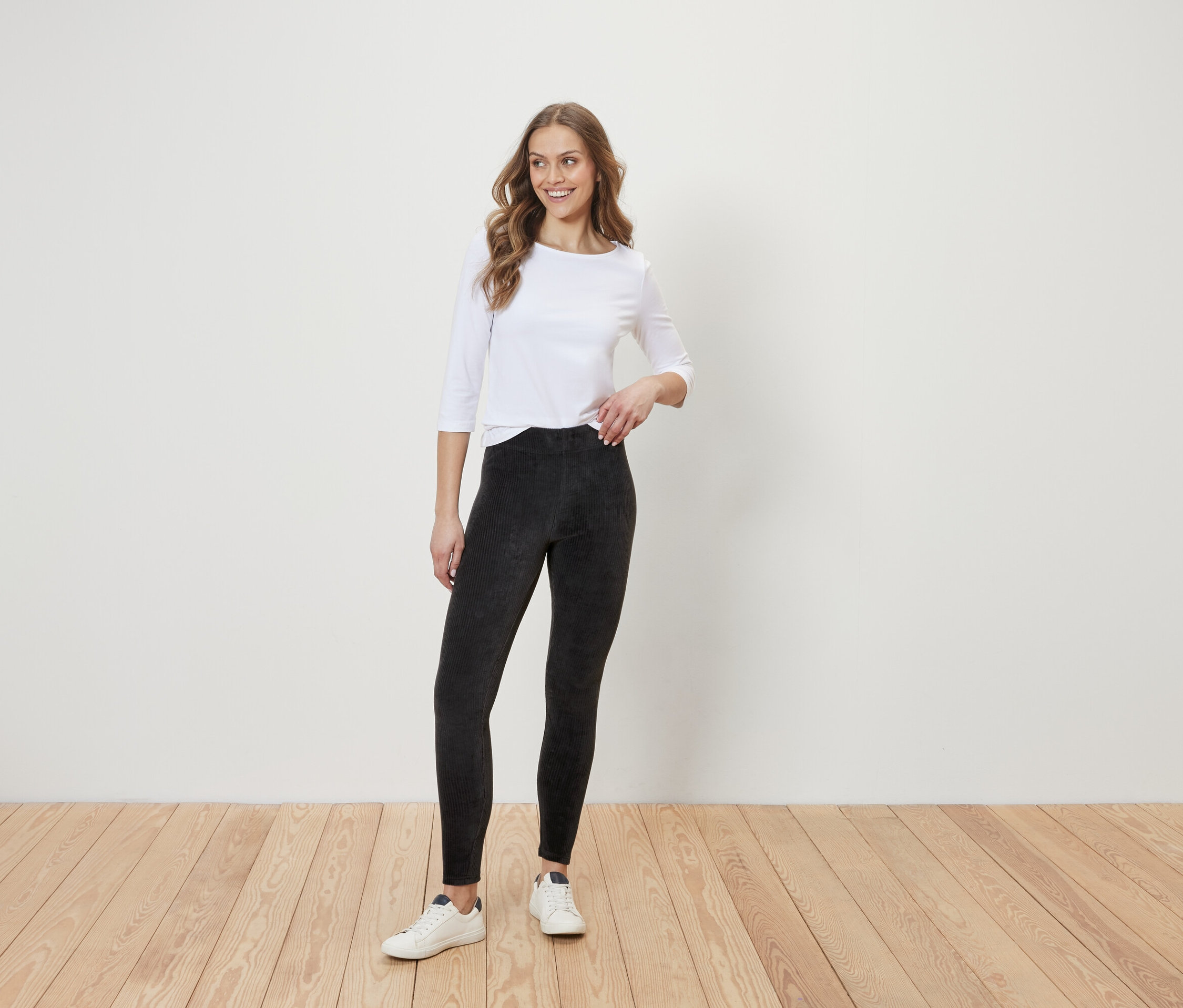 Eine Frau steht auf einem Holzboden vor einer weißen Wand. Sie trägt eine weiße Bluse mit Dreiviertelärmeln, schwarze Leggings und weiße Turnschuhe.