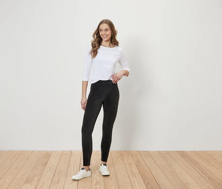 Eine Frau steht auf einem Holzboden vor einer weißen Wand. Sie trägt eine weiße Bluse mit Dreiviertelärmeln, schwarze Leggings und weiße Turnschuhe.