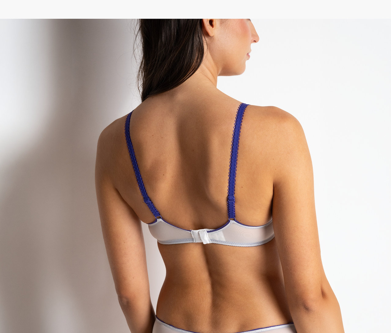 Rückenansicht einer Frau, die einen weißen Passionata Wired Plunge BH »Chloe« mit blauen Trägern und passenden Slips trägt.