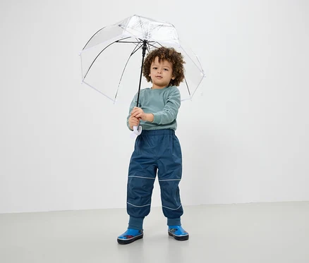 Ein Junge hält einen transparenten Regenschirm, trägt blaue Hosen und Gummistiefel.