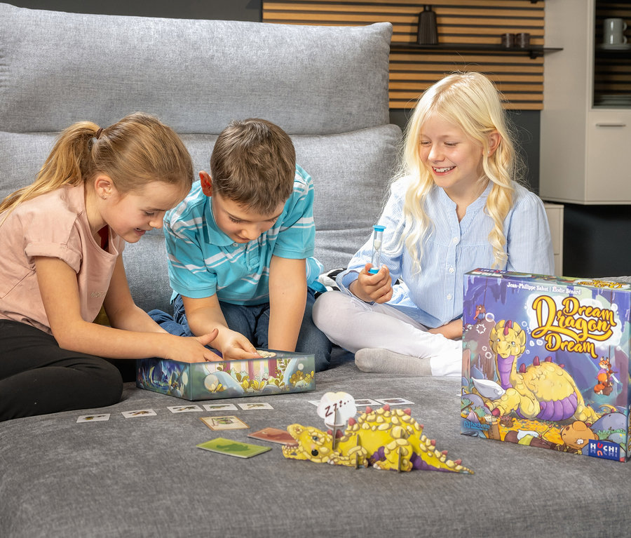 Drei Kinder spielen das Spiel »Dream Dragon Dream« auf einem grauen Sofa. Ein Kind hält eine Sanduhr.