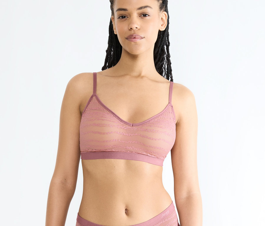 Eine Frau mit dunklen Zöpfen trägt einen pinkfarbenen sloggi FREE Evolve PU Lace BH und passende Spitzenunterwäsche vor weißem Hintergrund.