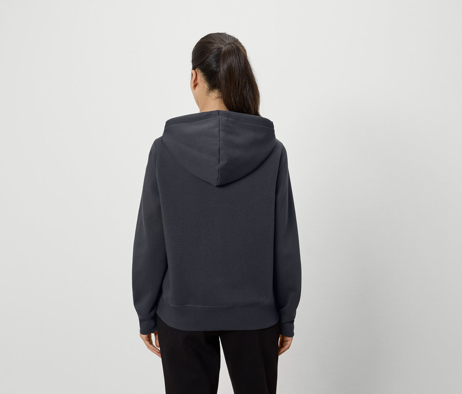 Eine Frau steht mit dem Rücken zur Kamera und trägt einen dunkelgrauen Kapuzenpullover und schwarze Hosen.