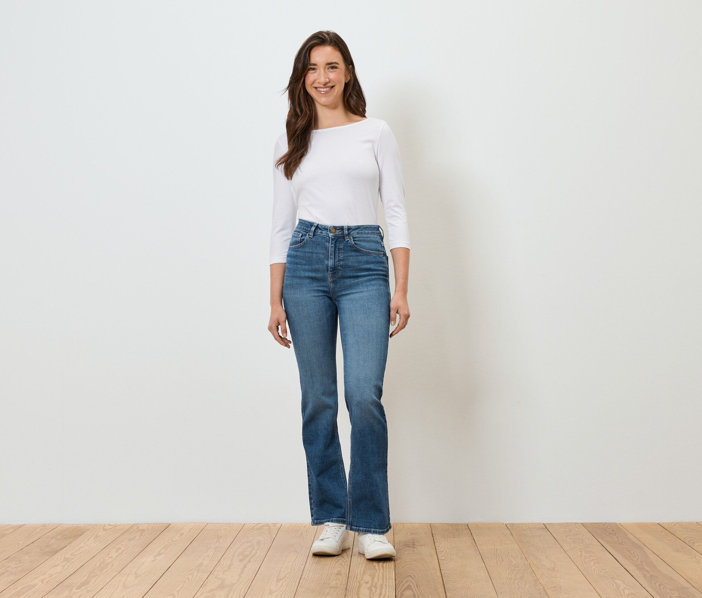 Eine Frau steht vor einer weißen Wand und trägt blaue Jeans, ein weißes Hemd und weiße Turnschuhe.