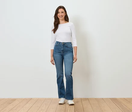 Eine Frau steht vor einer weißen Wand und trägt blaue Jeans, ein weißes Hemd und weiße Turnschuhe.