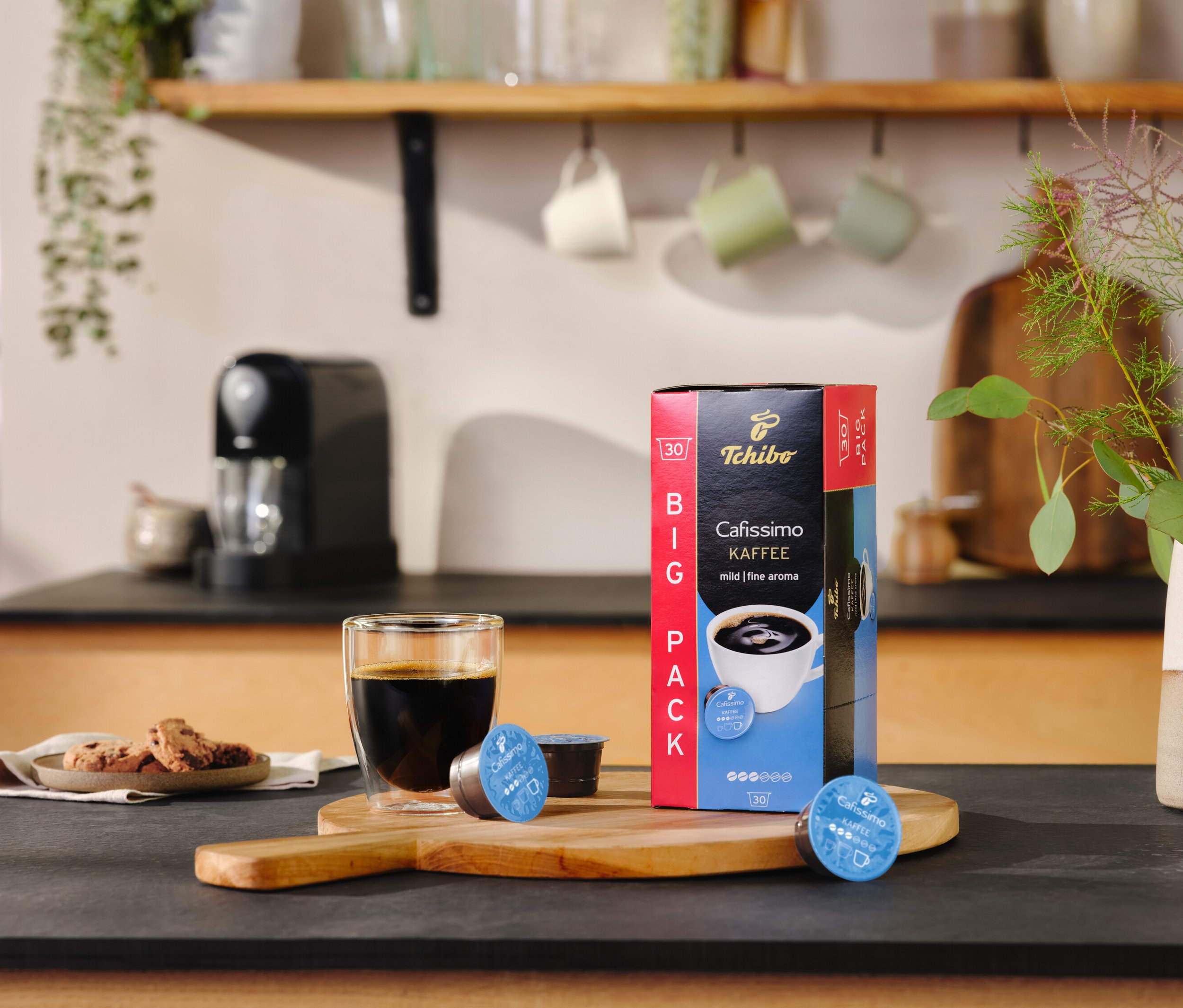 Auf einem Tisch stehen ein Glas Kaffee, eine Packung Cafissimo pure+, black und zwei Kapseln.