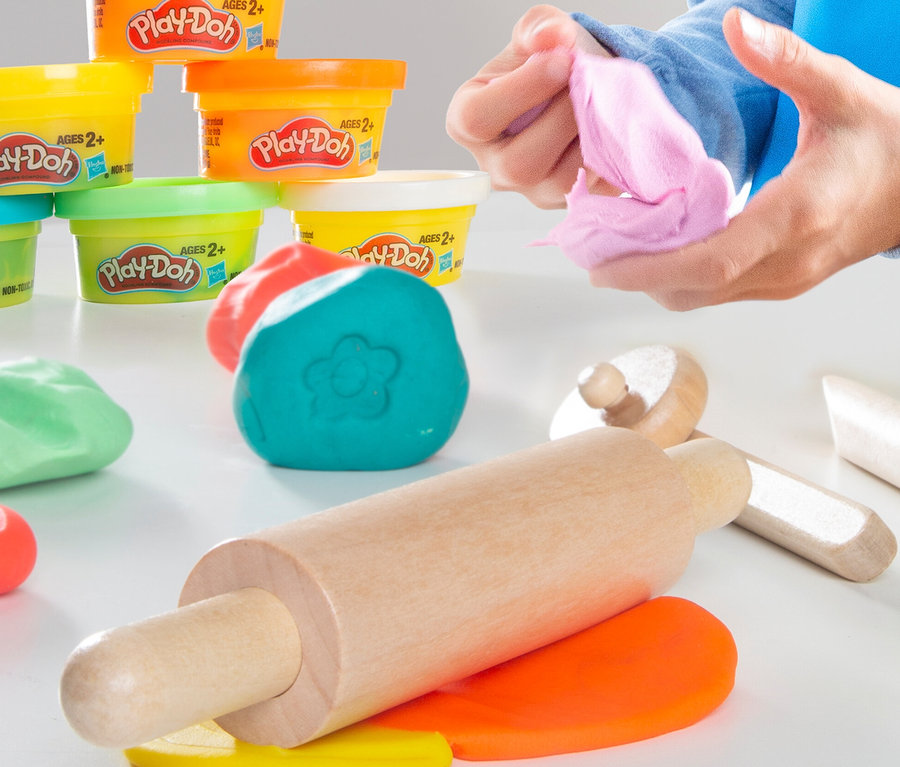 Person hält rosa Play-Doh Knete. Auf dem Tisch liegen eine Walze und weitere Farben des roba x Play-Doh Knet-Spielset 1.