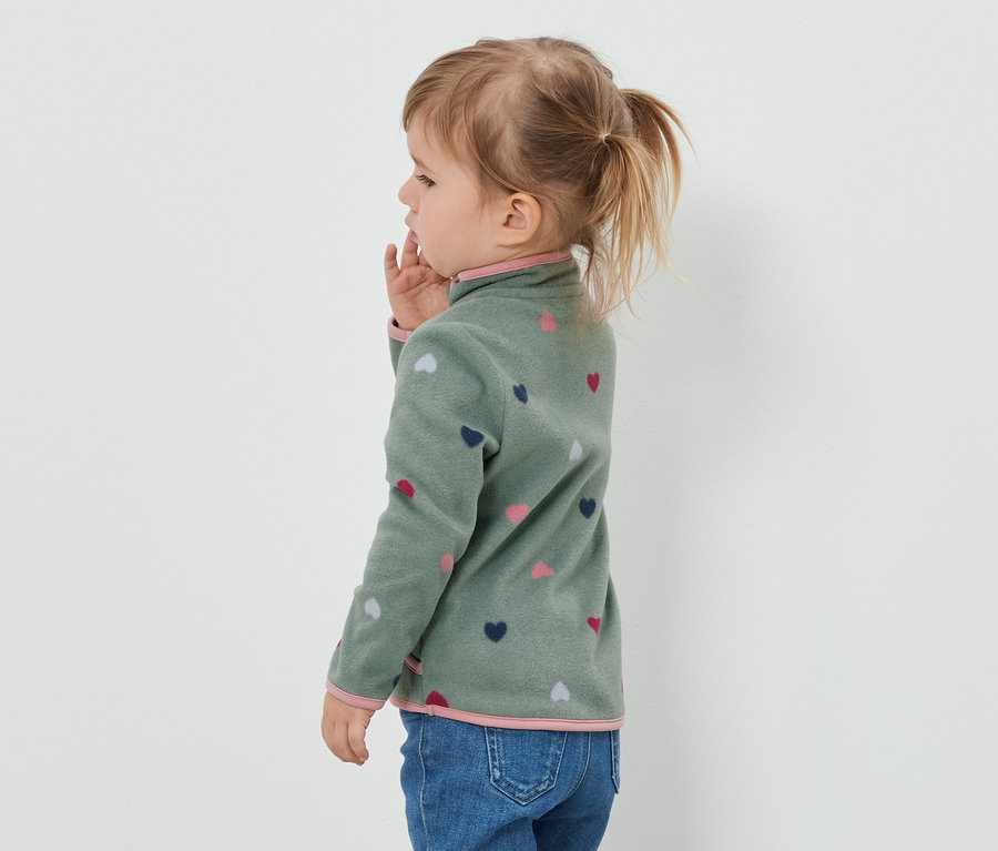 Seitenansicht eines kleinen Mädchens mit blonden, zum Pferdeschwanz gebundenen Haaren, das eine grüne Fleecejacke mit Herzmuster und Jeans trägt.