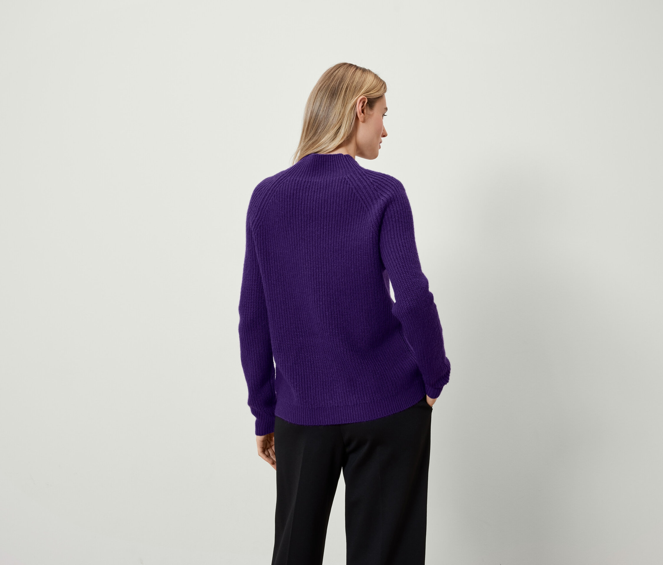 Frau mit dem Rücken zur Kamera trägt einen lila Pullover und eine schwarze Hose.