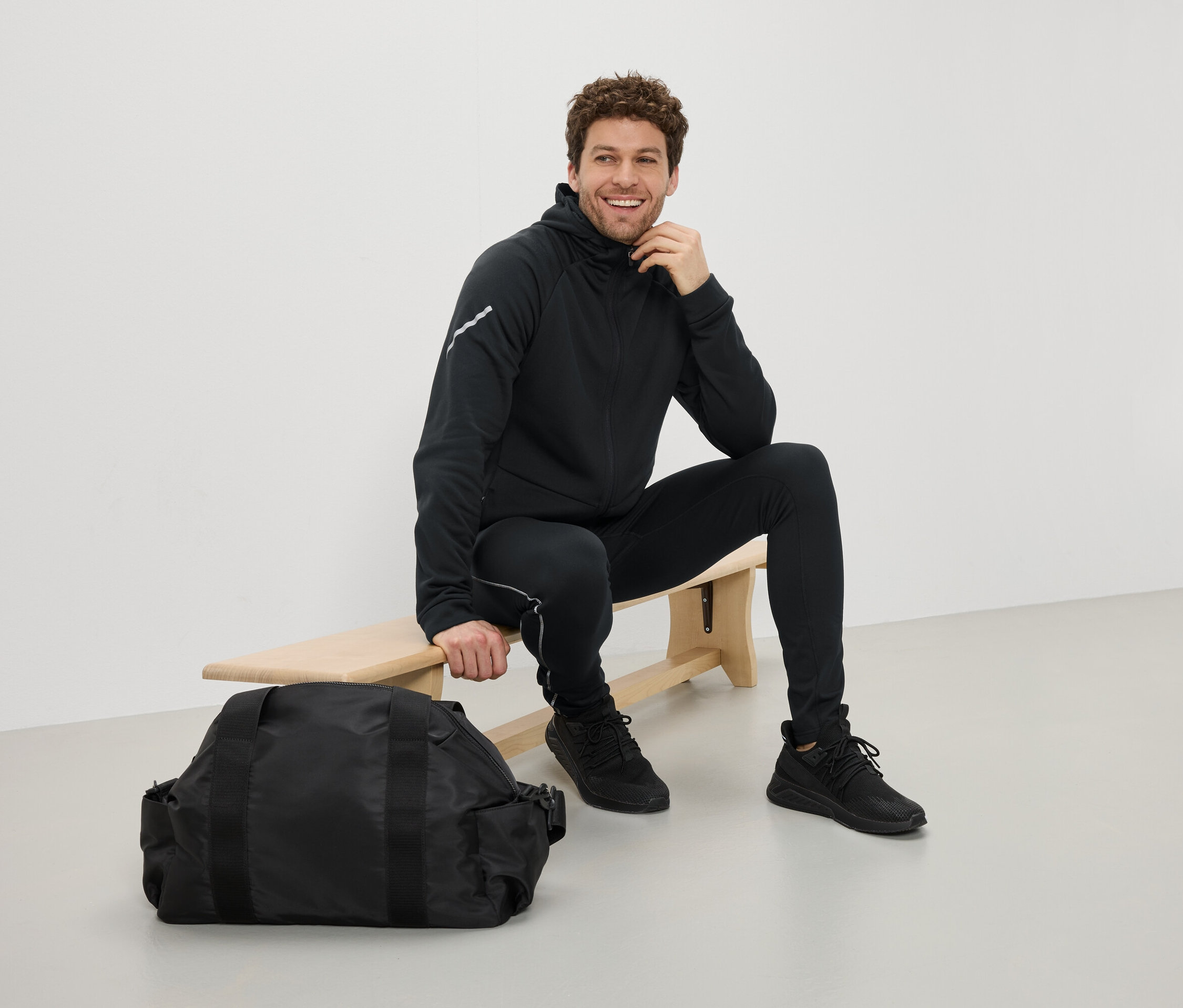 Mann sitzt in schwarzer Sportbekleidung auf einer Bank neben einer schwarzen Sporttasche.