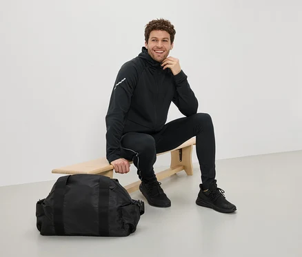 Mann sitzt in schwarzer Sportbekleidung auf einer Bank neben einer schwarzen Sporttasche.
