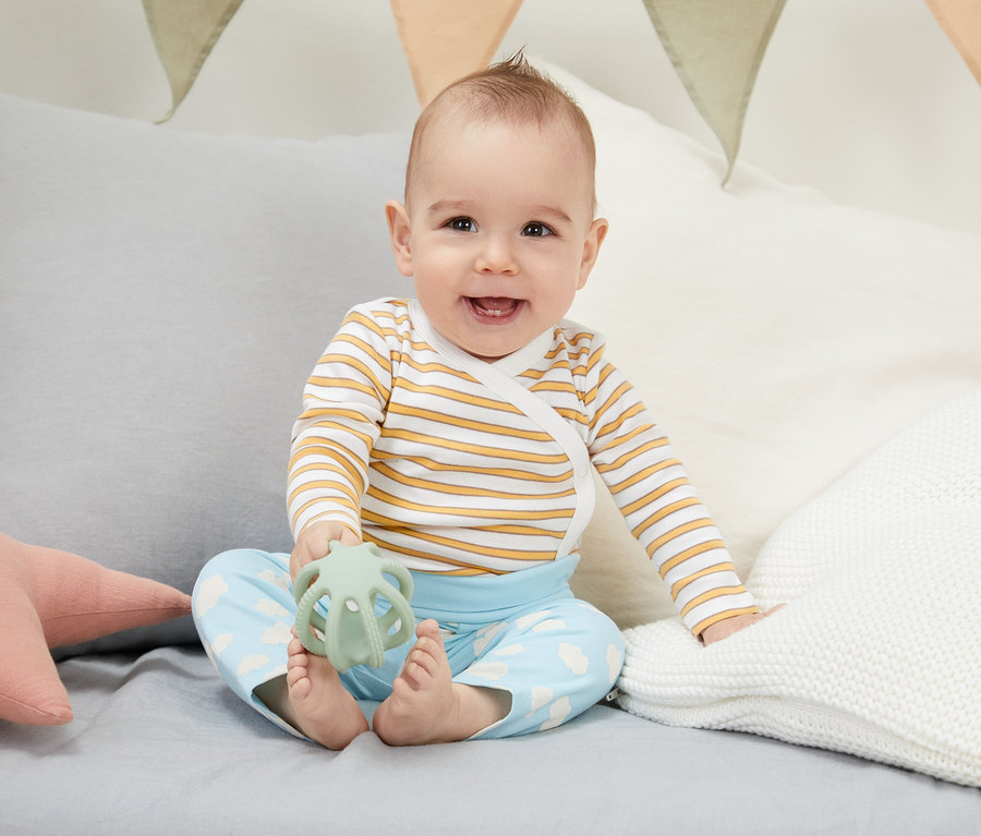 Ein lächelndes Baby sitzt auf einem Bett und hält ein grünes Spielzeug. Es trägt ein 4-teiliges Baby-Set aus Bio-Baumwolle.
